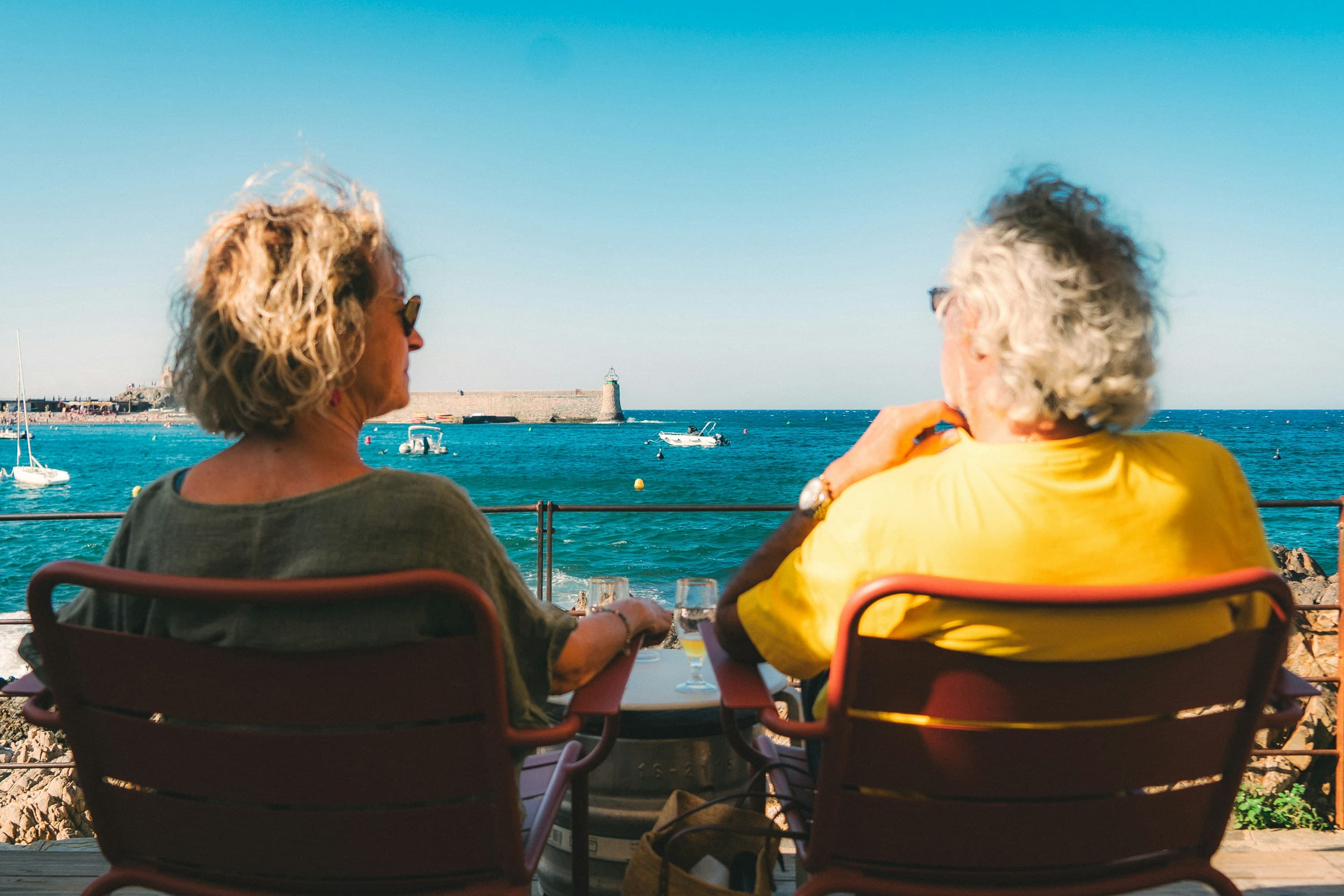 Two travellers sharing a glass of wine at a sunlit seaside cafe, laughing together as they look out over turquoise water