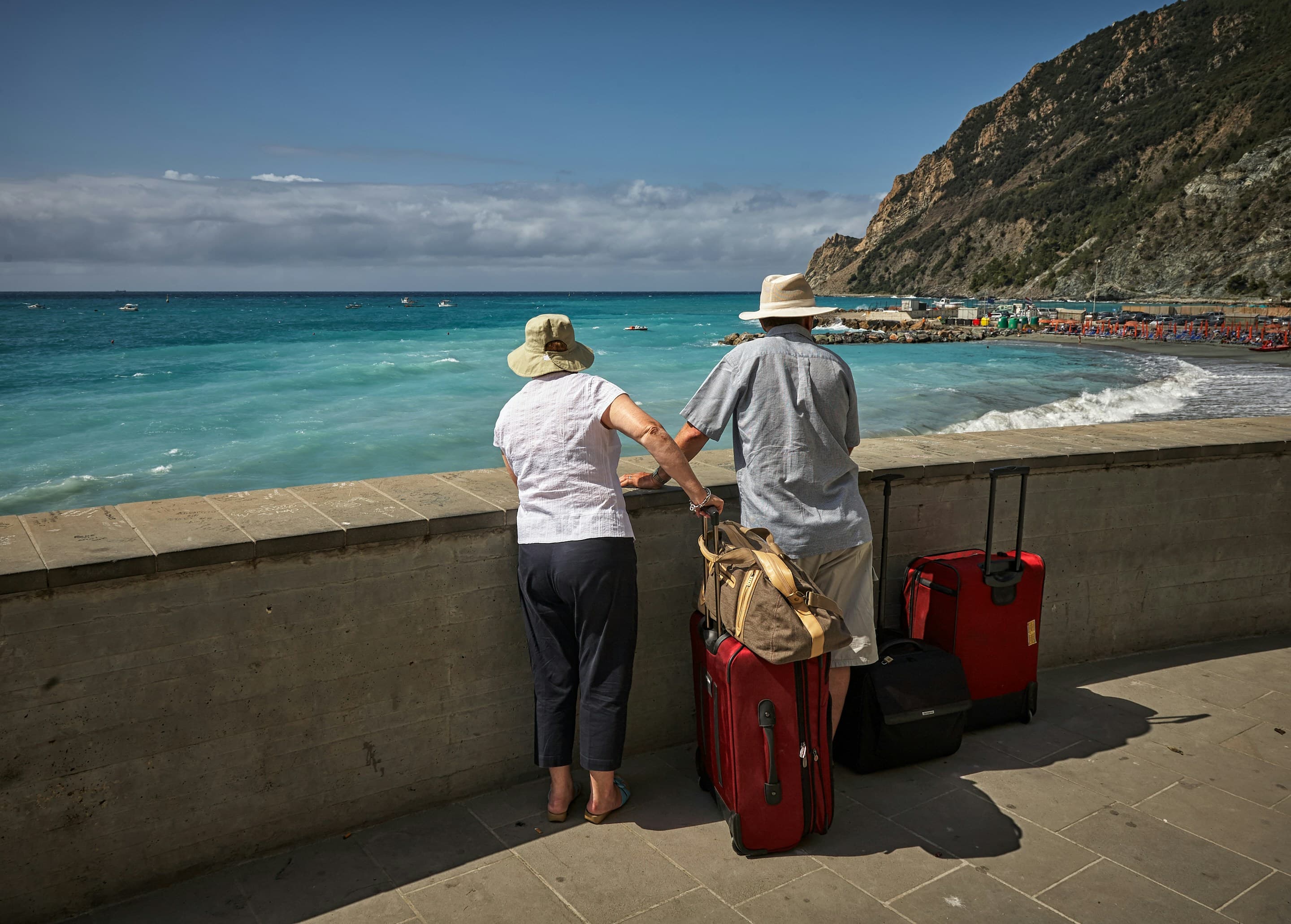 Two mature travellers pausing together at a seaside wall, taking in a scenic coastal view with their luggage beside them